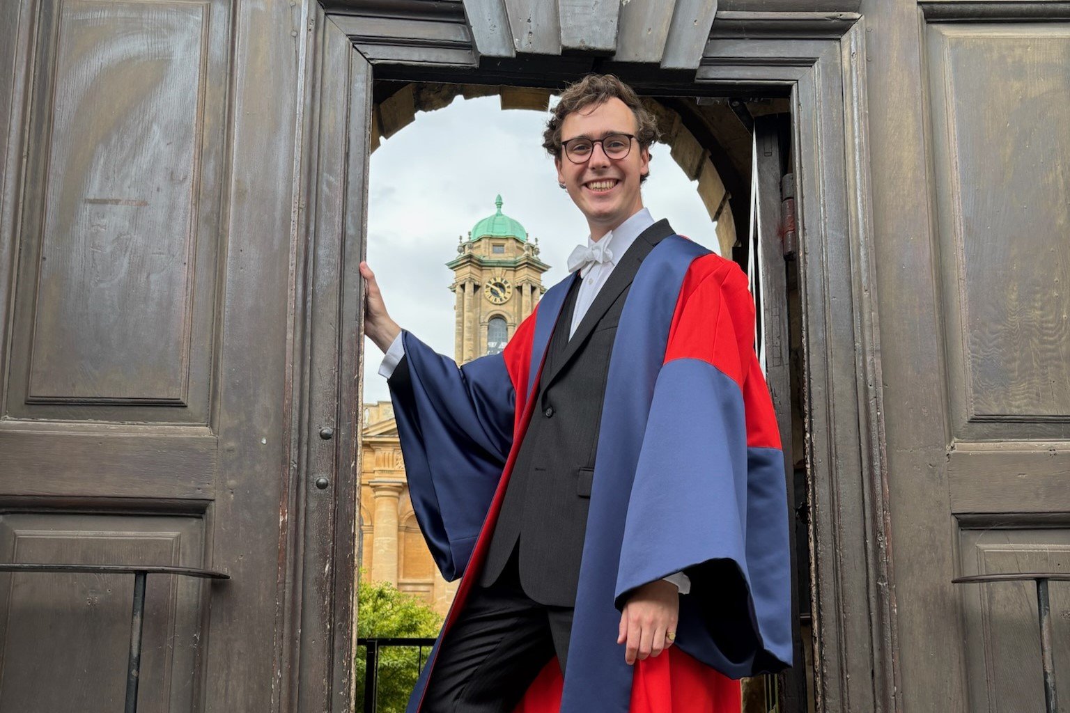 Samuel Teague standing in the the doorway of The Queen's College, Oxford, wearing doctoral (DPhil) robes.